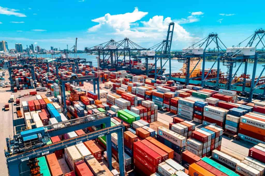 A busy port terminal filled with colorful shipping containers and large industrial cranes, illustrating how vessel schedule changes are leading to global cargo shipping disruptions in 2026.